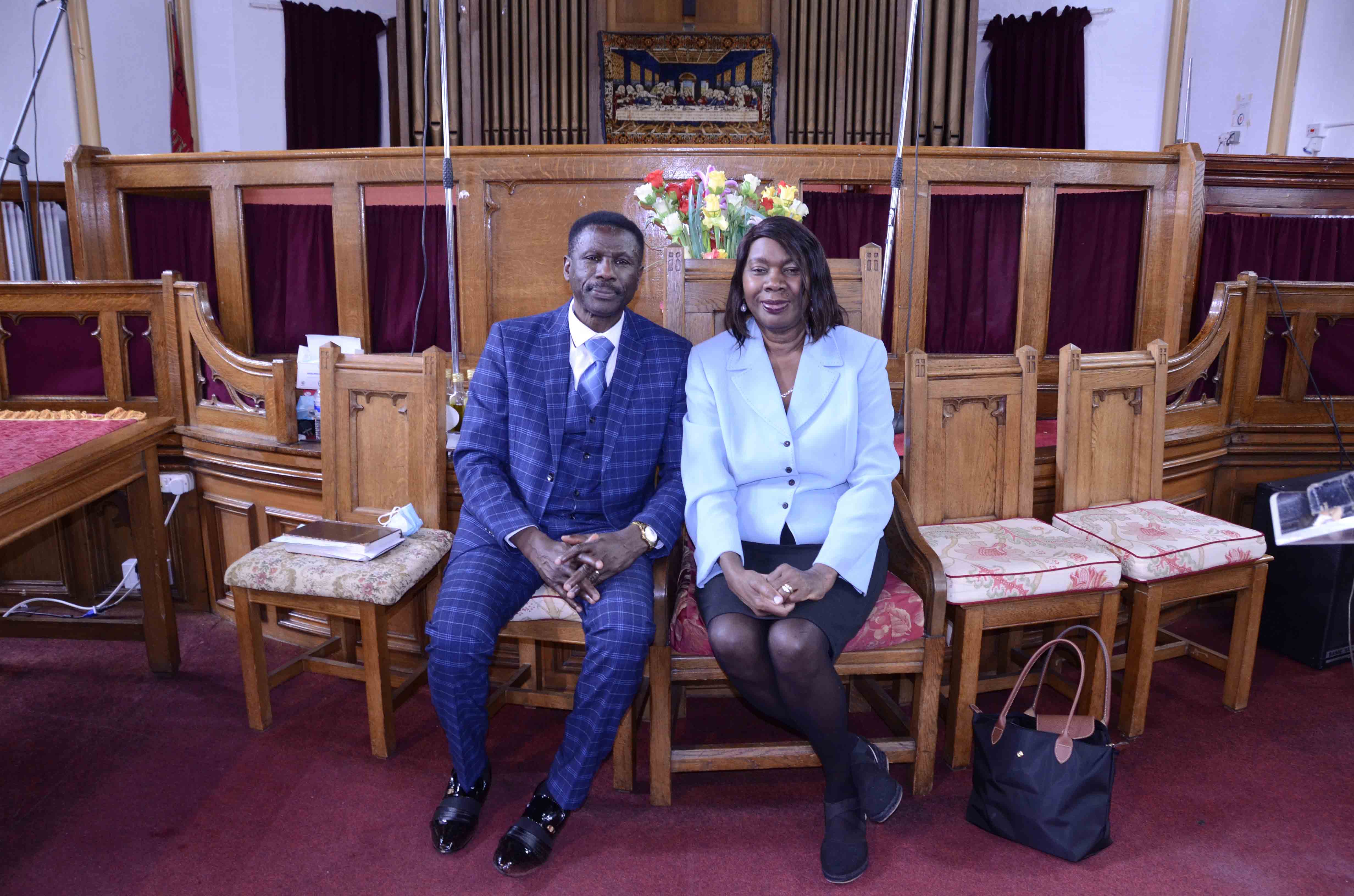Pastor and Mrs Mereweather-Thompson seated at the front of the modern sanctuary, with the Last Supper tapestry visible behind them and the baptism pool hidden behind the new bench